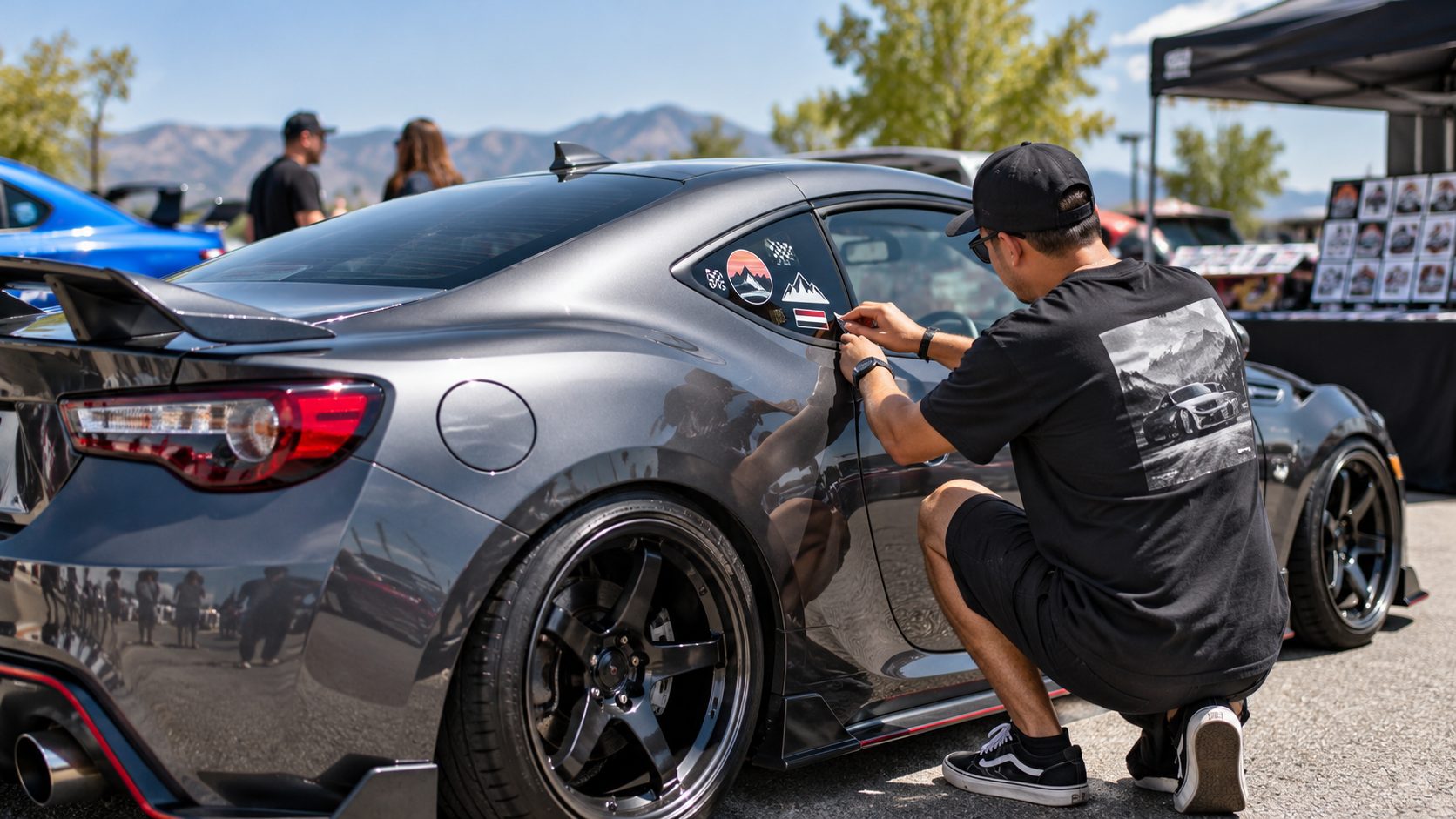 Man applying decorative vinyl stickers to the rear side window of a grey sports car at an outdoor automotive event.
